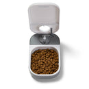 A grey and white automatic pet feeder for a cat or a small dog with dry pet food in it, on a white background