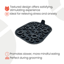 Infographic of a black textured pet licking mat with raised circular patterns and a small handle tab. Text highlights benefits including: textured design offers a satisfying, stimulating experience; ideal for relieving stress and anxiety; promotes slower, more mindful eating; and is perfect for use during grooming. Accompanied by small icons representing a cat face, a medical cross, a clock, and grooming tools.