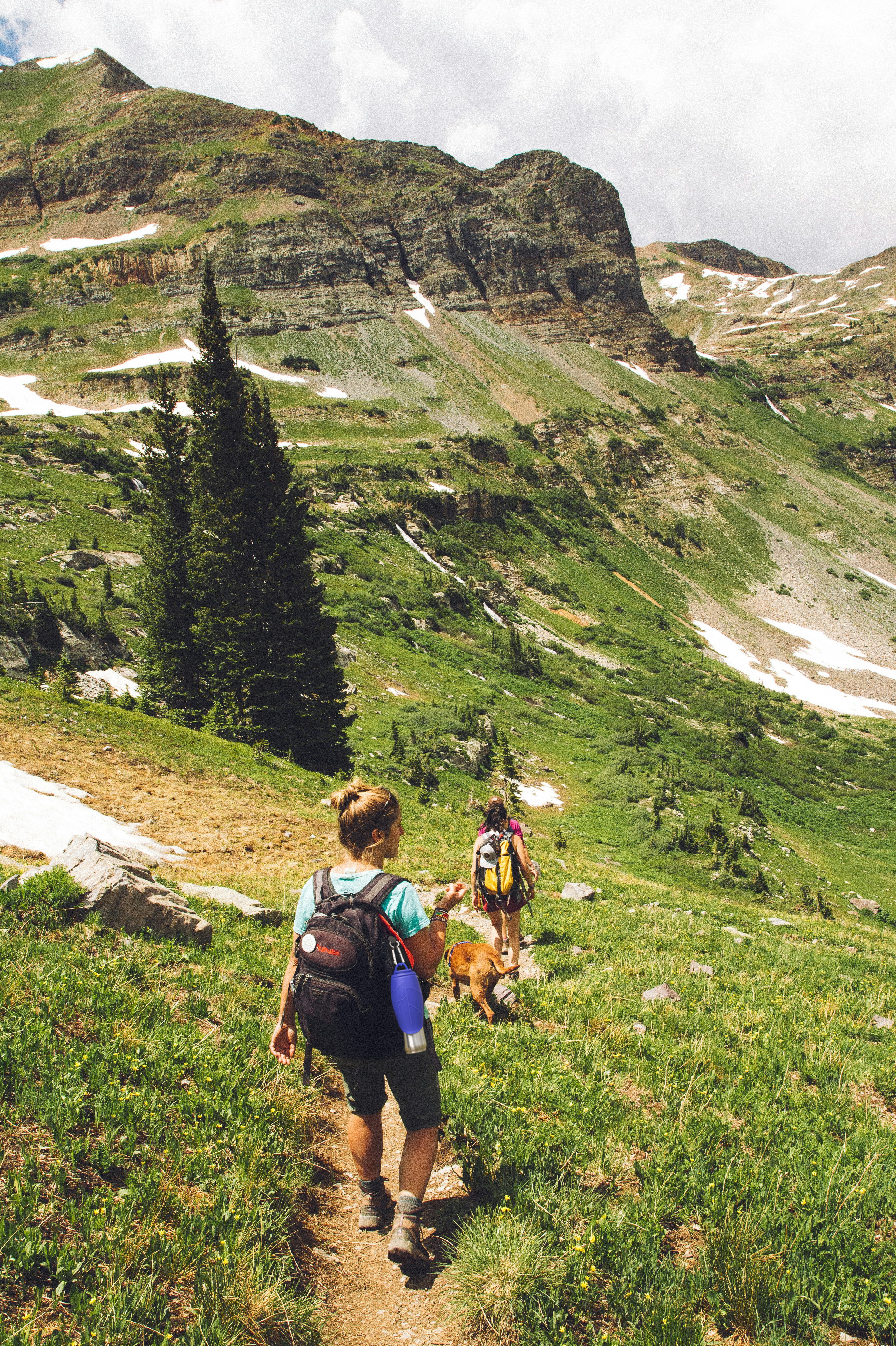 Two hikers with backpacks walking on a trail in a mountainous area with green grass and rocky terrain, with the Dog Mate insulated flask clipped to the dark coloured backpack of the hiker in the foreground. 